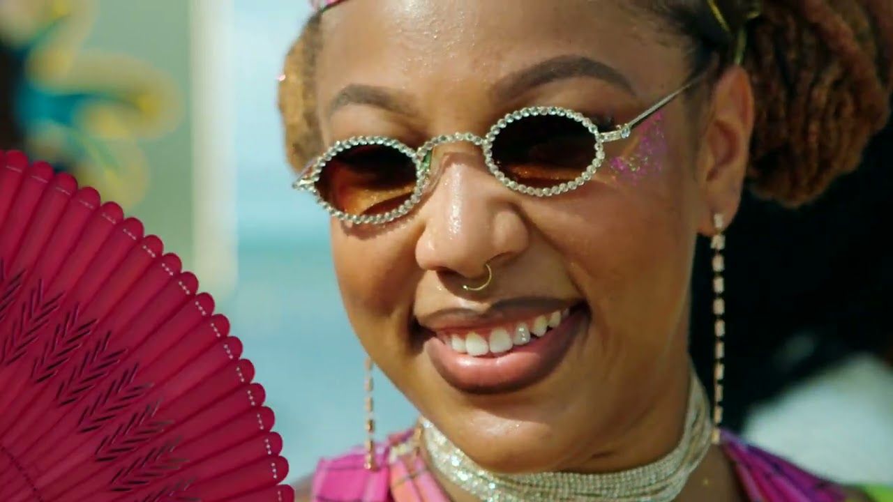 Gros plan d'une festivalière souriante, lunettes rondes serties et paillettes sur la tempe, un éventail rose au premier plan.